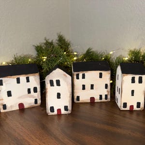 May include: Four small, handcrafted wooden houses with white walls, black roofs, and black window details. Each house has a small red door. The houses are arranged on a wooden surface, with greenery and fairy lights in the background.