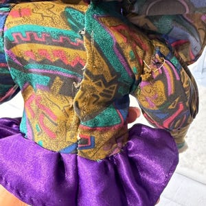 K&ouml;nnte beinhalten: Eine Vintage-Puppe mit einem gemusterten Stoffkleid in Braun-, T&uuml;rkis-, Lila- und Rosat&ouml;nen. Das Kleid der Puppe hat einen ger&uuml;schten lila Rock. Die Arme und der Kopf der Puppe sind sichtbar.