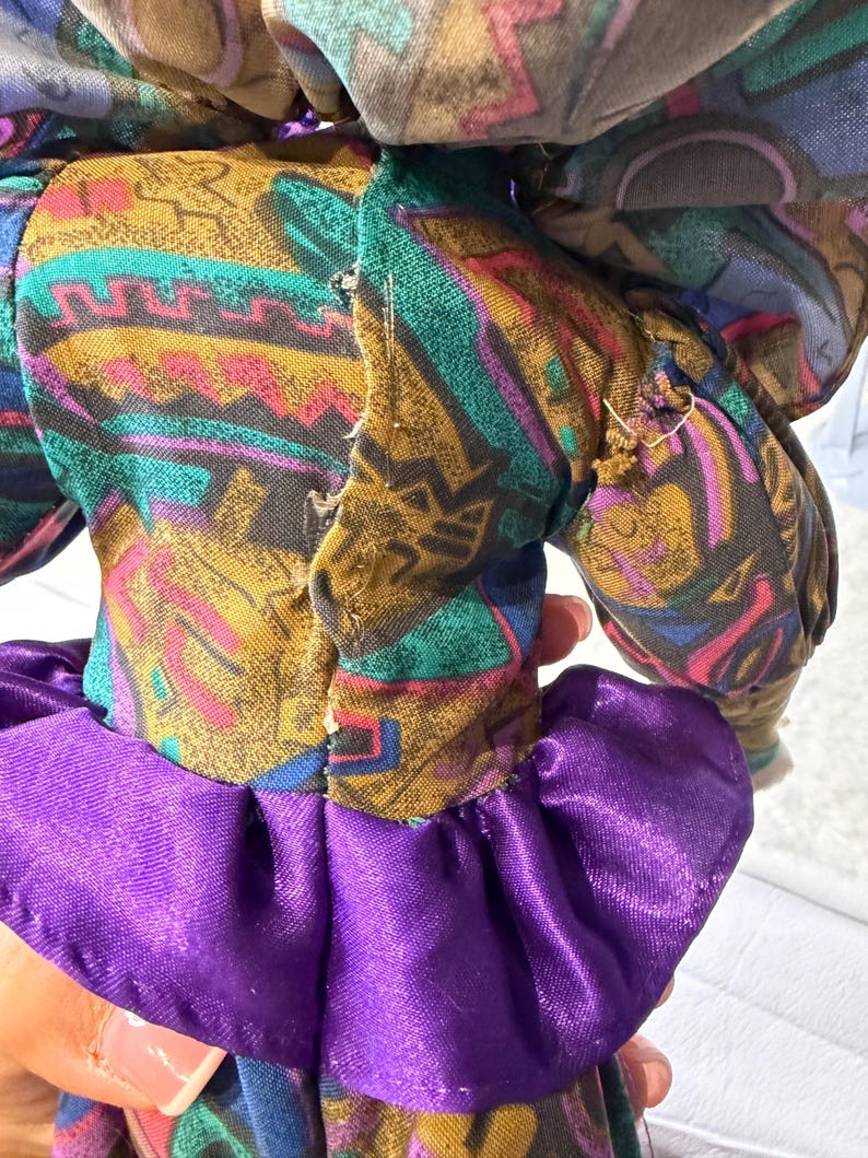 K&ouml;nnte beinhalten: Eine Vintage-Puppe mit einem gemusterten Stoffkleid in Braun-, T&uuml;rkis-, Lila- und Rosat&ouml;nen. Das Kleid der Puppe hat einen ger&uuml;schten lila Rock. Die Arme und der Kopf der Puppe sind sichtbar.