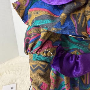 K&ouml;nnte beinhalten: Eine Vintage-Puppe in einem farbenfrohen Kleid mit geometrischen Mustern in Blau, Gold, Rosa und T&uuml;rkis. Das Kleid hat einen lila Satin-Kragen und Taillenbesatz. Der &Auml;rmel der Puppe hat eine ger&uuml;schte Manschette.