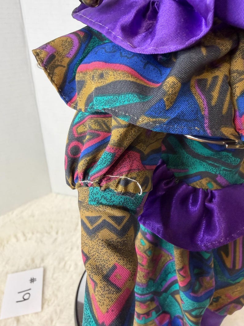 K&ouml;nnte beinhalten: Eine Vintage-Puppe in einem farbenfrohen Kleid mit geometrischen Mustern in Blau, Gold, Rosa und T&uuml;rkis. Das Kleid hat einen lila Satin-Kragen und Taillenbesatz. Der &Auml;rmel der Puppe hat eine ger&uuml;schte Manschette.