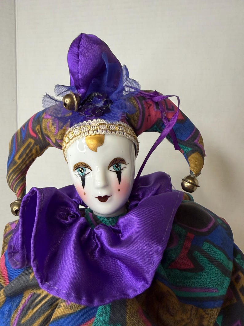 K&ouml;nnte beinhalten: Dekorative Porzellan-Narrenpuppe mit wei&szlig;em Gesicht, blauen Augen und rotem Rouge. Die Puppe tr&auml;gt einen lila Hut mit goldenen Gl&ouml;ckchen und einen passenden Kragen. Das Kost&uuml;m des Narren hat einen bunten Stoff mit Muster.