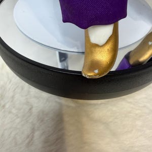 K&ouml;nnte beinhalten: Nahaufnahme eines goldfarbenen Schuhs einer Figur mit wei&szlig;em Akzent und lila Stoff. Die Figur steht auf einer wei&szlig;en kreisf&ouml;rmigen Basis mit schwarzem Rand. Der Schuh hat eine kleine Stelle, an der die Goldfarbe abgebl&auml;ttert ist.