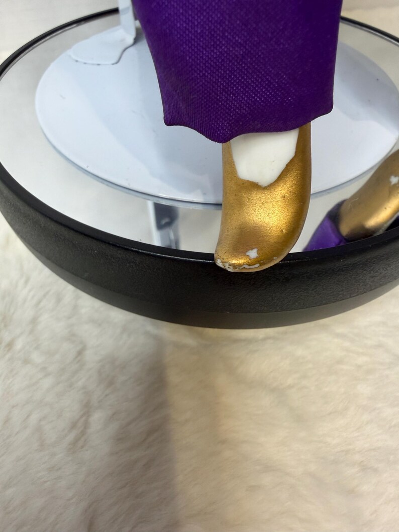 K&ouml;nnte beinhalten: Nahaufnahme eines goldfarbenen Schuhs einer Figur mit wei&szlig;em Akzent und lila Stoff. Die Figur steht auf einer wei&szlig;en kreisf&ouml;rmigen Basis mit schwarzem Rand. Der Schuh hat eine kleine Stelle, an der die Goldfarbe abgebl&auml;ttert ist.