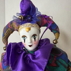 K&ouml;nnte beinhalten: Dekorative Porzellan-Narrenpuppe mit wei&szlig;em Gesicht, blauen Augen und roten Lippen. Die Puppe tr&auml;gt einen lila Satin-Kragen und ein gemustertes Stoff-Outfit mit passendem lila Hut, der mit Glocken verziert ist. Der Hut hat eine goldene Verzierung.