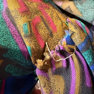 K&ouml;nnte beinhalten: Nahaufnahme eines Stoffes mit einem lebendigen, mehrfarbigen Muster. Das Design zeigt Streifen in Gold, Pink, Lila und T&uuml;rkis. Der Stoff scheint gewebt zu sein, mit sichtbaren F&auml;den und einem leicht ausgefransten Rand. Die Gesamt&auml;sthetik ist k&uuml;hn und farbenfroh.
