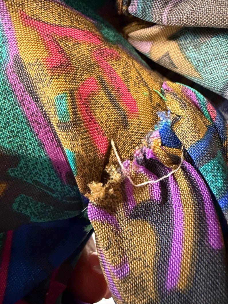 K&ouml;nnte beinhalten: Nahaufnahme eines Stoffes mit einem lebendigen, mehrfarbigen Muster. Das Design zeigt Streifen in Gold, Pink, Lila und T&uuml;rkis. Der Stoff scheint gewebt zu sein, mit sichtbaren F&auml;den und einem leicht ausgefransten Rand. Die Gesamt&auml;sthetik ist k&uuml;hn und farbenfroh.