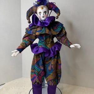K&ouml;nnte beinhalten: Porzellan-Narrenpuppe mit wei&szlig;em Gesicht, schwarzen tr&auml;nenf&ouml;rmigen Markierungen und einem lila und gemusterten Kost&uuml;m. Die Puppe hat einen ger&uuml;schten Kragen, einen passenden Hut mit Gl&ouml;ckchen und goldene Schuhe. Die Puppe steht auf einem schwarzen St&auml;nder.