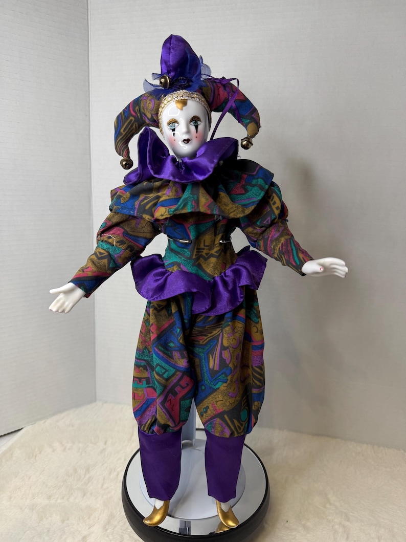 K&ouml;nnte beinhalten: Porzellan-Narrenpuppe mit wei&szlig;em Gesicht, schwarzen tr&auml;nenf&ouml;rmigen Markierungen und einem lila und gemusterten Kost&uuml;m. Die Puppe hat einen ger&uuml;schten Kragen, einen passenden Hut mit Gl&ouml;ckchen und goldene Schuhe. Die Puppe steht auf einem schwarzen St&auml;nder.