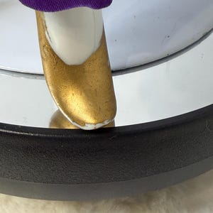 K&ouml;nnte beinhalten: Nahaufnahme eines Puppenfu&szlig;es mit einem goldfarbenen Schuh. Der Schuh befindet sich auf einem wei&szlig;en Fu&szlig; und ist mit einem lila Kleidungsst&uuml;ck kombiniert. Die Puppe befindet sich auf einer reflektierenden Oberfl&auml;che.