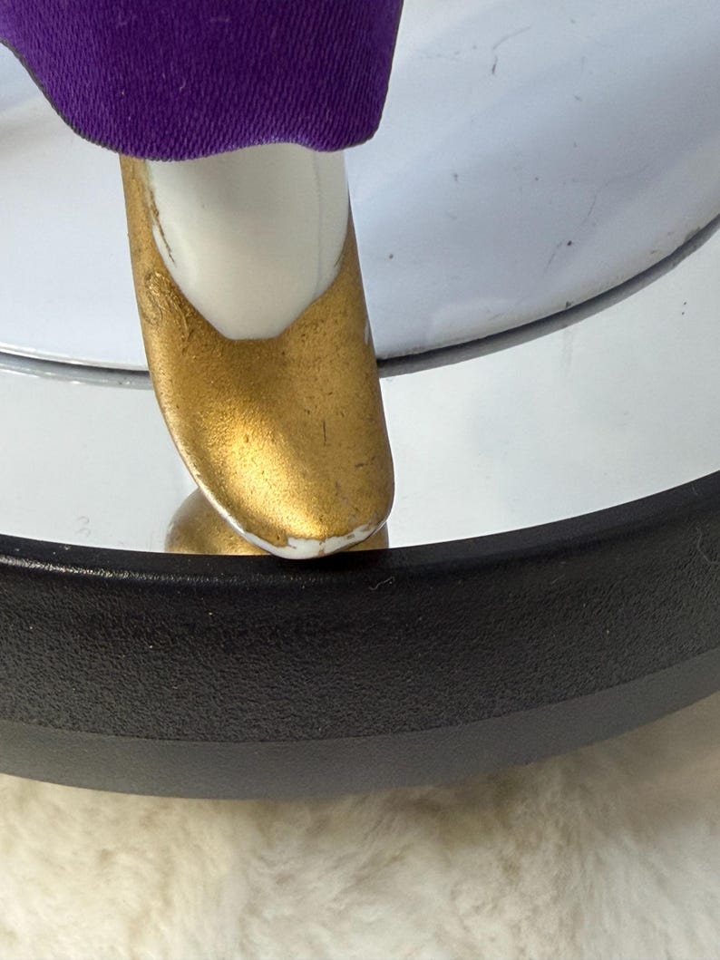 K&ouml;nnte beinhalten: Nahaufnahme eines Puppenfu&szlig;es mit einem goldfarbenen Schuh. Der Schuh befindet sich auf einem wei&szlig;en Fu&szlig; und ist mit einem lila Kleidungsst&uuml;ck kombiniert. Die Puppe befindet sich auf einer reflektierenden Oberfl&auml;che.
