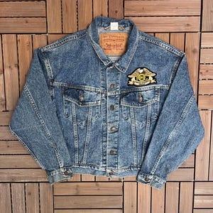 May include: A vintage Levi Strauss & Co. denim jacket in a medium wash. The jacket features a Harley Owners Group patch on the chest and a Levi's label on the collar. It has two chest pockets and button closures. The jacket is laid flat on a wooden surface.