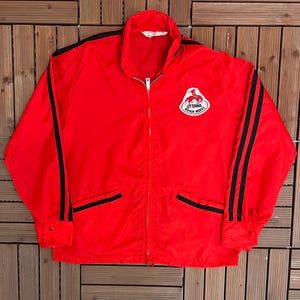 May include: A red zip-up jacket with black stripes on the sleeves and a logo patch on the chest. The jacket has two front pockets and a stand-up collar. The logo patch reads "Ottawa Rough Riders".