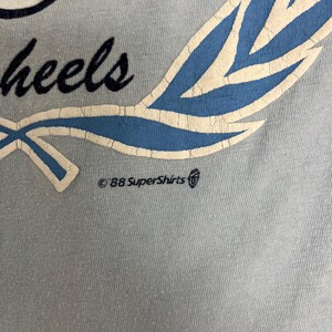 May include: Close-up of a light blue t-shirt with a design featuring the word "wheels" in a script font, a blue and white laurel wreath, and the text "&copy;88 SuperShirts". The shirt has a vintage aesthetic.