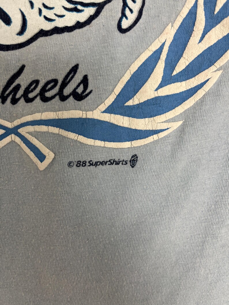 May include: Close-up of a light blue t-shirt with a design featuring the word "wheels" in a script font, a blue and white laurel wreath, and the text "&copy;88 SuperShirts". The shirt has a vintage aesthetic.