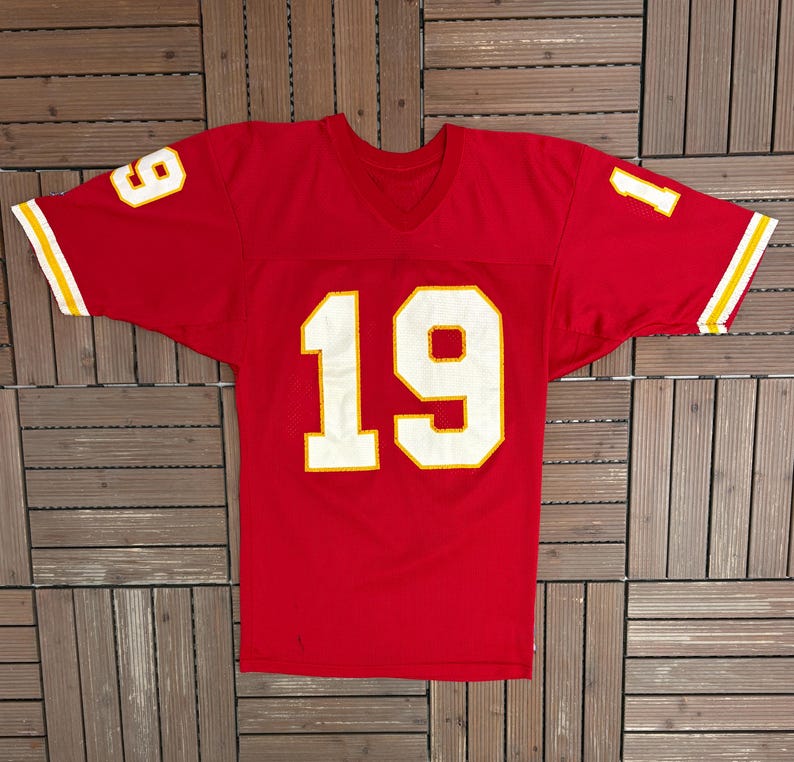 May include: A red football jersey featuring the number 19 in white with a yellow outline on the front. The number 9 is on the left shoulder and the number 1 on the right. Sleeves have yellow and white stripes. A classic sports jersey.