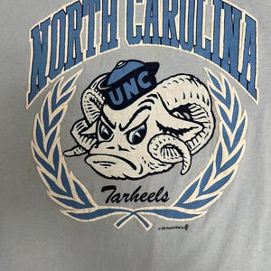 May include: Light blue t-shirt featuring a distressed graphic of a ram wearing a UNC hat, surrounded by a laurel wreath. The text "North Carolina" arches across the top, with "Tarheels" below.