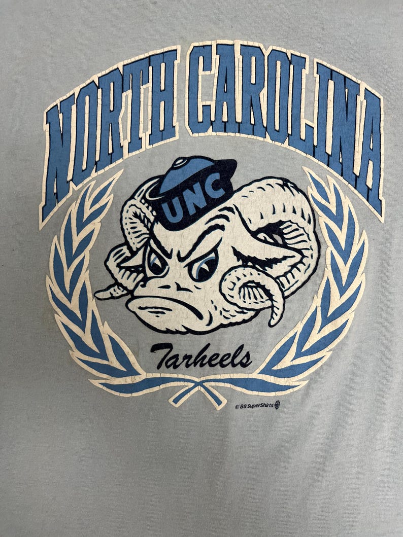 May include: Light blue t-shirt featuring a distressed graphic of a ram wearing a UNC hat, surrounded by a laurel wreath. The text "North Carolina" arches across the top, with "Tarheels" below.