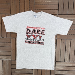 May include: A light gray Gildan Ultra Blend t-shirt with the text "PROPERTY OF D.A.R.E. XXL PROGRAM" in red and black. The shirt is laid flat on a wooden surface.