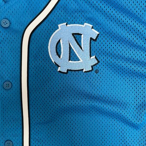 May include: Close-up of a light blue baseball jersey with a white and black trim. The jersey features the embroidered interlocking "NC" logo in light blue and white. The jersey has a button-up front.