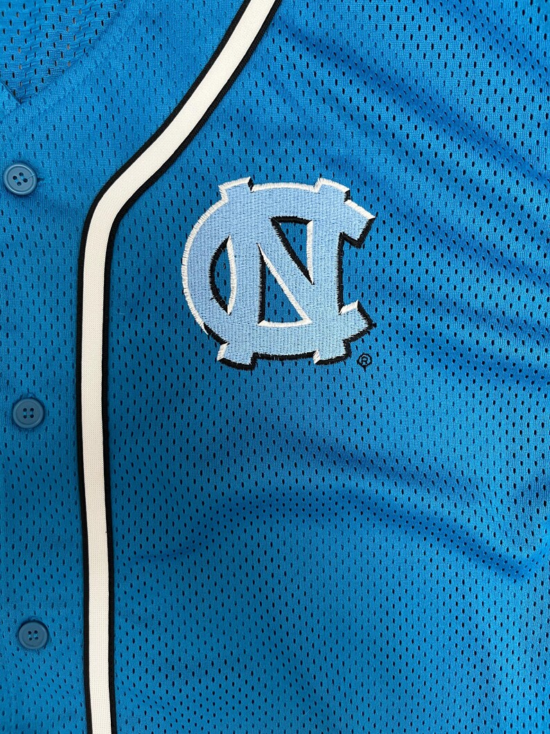 May include: Close-up of a light blue baseball jersey with a white and black trim. The jersey features the embroidered interlocking "NC" logo in light blue and white. The jersey has a button-up front.