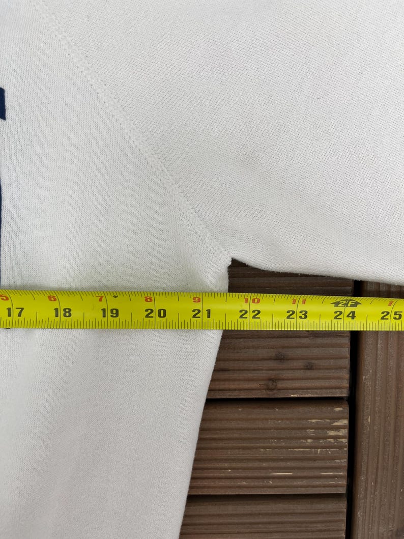 K&ouml;nnte beinhalten: Ein wei&szlig;es Sweatshirt mit dunkelblauem Akzent. Ein gelbes Ma&szlig;band zeigt eine Brustweite von ca. 61 cm. Das Sweatshirt ist aus einem weichen, strukturierten Stoff gefertigt.