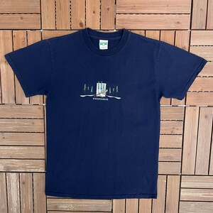 Pode incluir: Camiseta azul marinho com um desenho bordado de um edifício e árvores, com a palavra "KANANASKIS" abaixo. A camiseta tem mangas curtas e gola redonda.