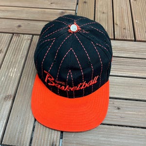Könnte beinhalten: Eine schwarze Baseballkappe mit roten, gestickten Nadelstreifen, die von einem silbernen Knopf in der Mitte ausgehen. Auf der Vorderseite befindet sich der rote Stickschriftzug Nike Basketball. Die Kappe hat einen flachen, orangefarbenen Schirm.