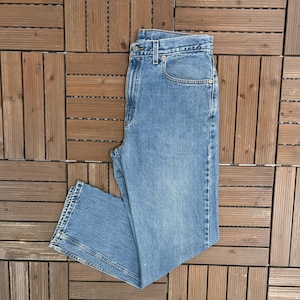 May include: A pair of light blue denim jeans, with a classic five-pocket design. The jeans are laid flat, showing the front and part of the back. The denim has a medium wash, with visible stitching and a button and zip closure. The jeans are a standard fit.