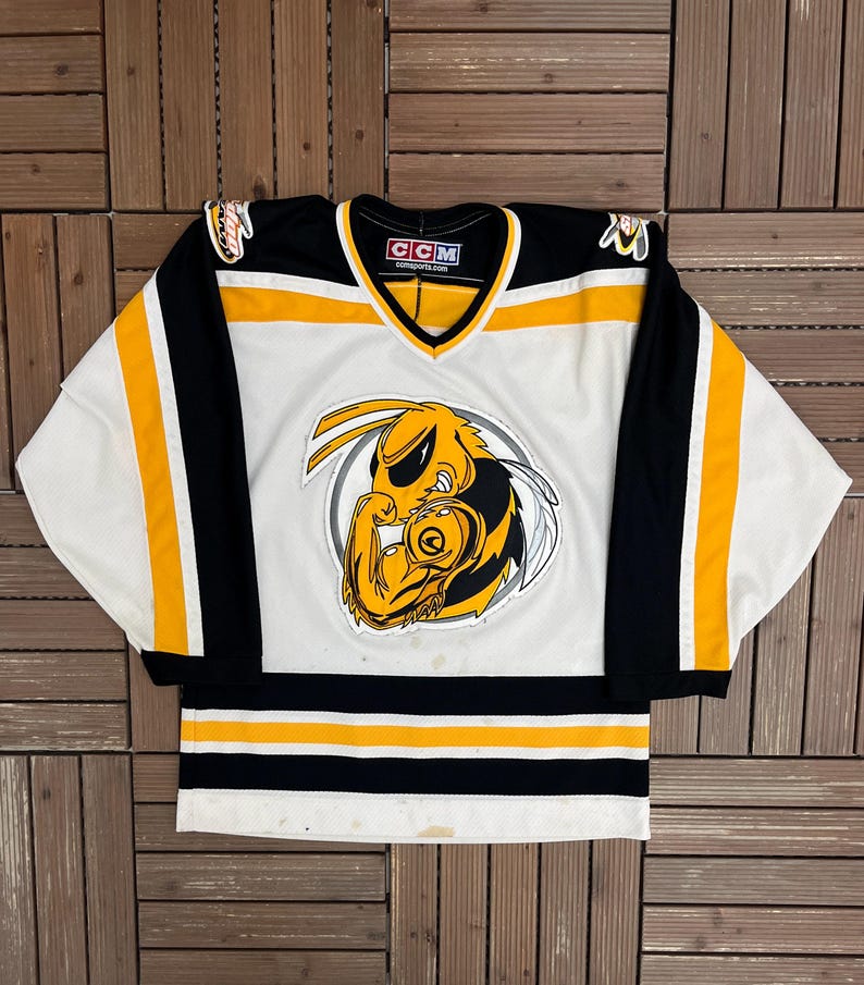 May include: White, black, and yellow hockey jersey with a V-neck collar. The jersey features a large cartoon wasp emblem on the front. The sleeves are black with yellow stripes. The jersey has a CCM tag.