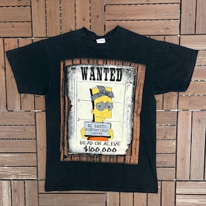 May include: Black t-shirt featuring a "Wanted" poster design with a cartoon character. The poster includes the text "El Barto Winterfield 159736" and "Dead or Alive $100,000". The design is framed in a wooden border.