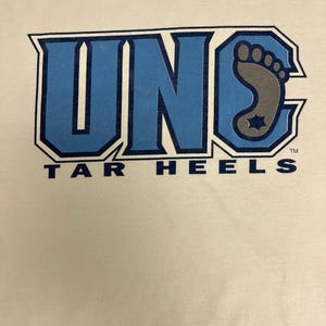 May include: Cream-coloured t-shirt with a blue and grey graphic. The graphic displays the letters "UNC" in blue, outlined in a darker blue, with a grey foot design and the words "TAR HEELS" in blue.