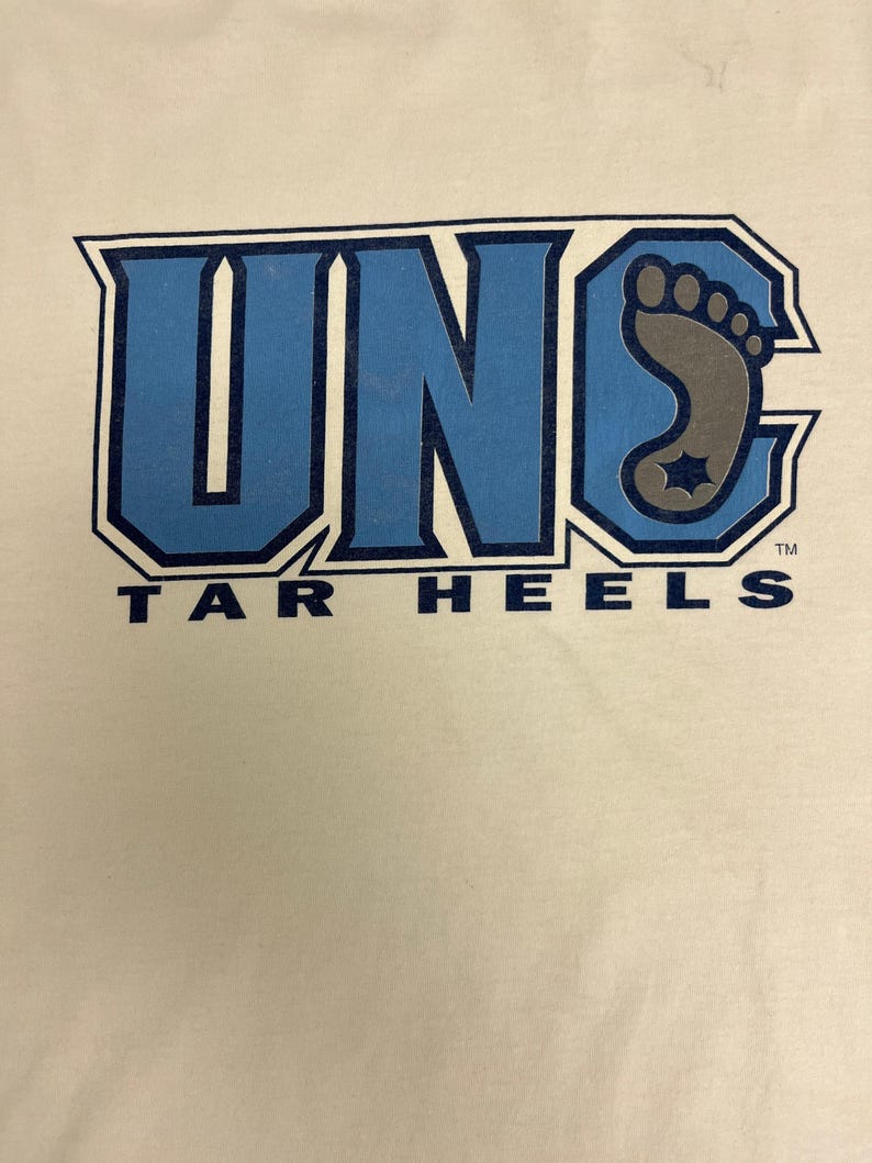 May include: Cream-coloured t-shirt with a blue and grey graphic. The graphic displays the letters "UNC" in blue, outlined in a darker blue, with a grey foot design and the words "TAR HEELS" in blue.