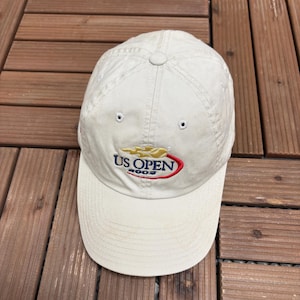 Op de afbeelding: Beige baseballpet met de tekst "US OPEN 2003" geborduurd in rood, goud en blauw. De pet heeft een gebogen klep en een ronde kroon met ventilatiegaten.