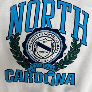 K&ouml;nnte beinhalten: Wei&szlig;es Sweatshirt mit den Worten "NORTH CAROLINA" in blauen Blockbuchstaben. In der Mitte befindet sich ein rundes Emblem mit dem Text "UNIVERSITY OF NORTH CAROLINA CHAPEL HILL, N.C." und dem Wort "TARHEELS".