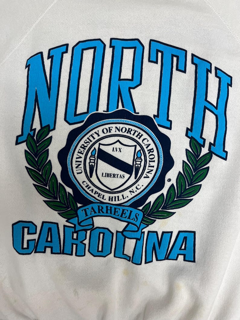 K&ouml;nnte beinhalten: Wei&szlig;es Sweatshirt mit den Worten "NORTH CAROLINA" in blauen Blockbuchstaben. In der Mitte befindet sich ein rundes Emblem mit dem Text "UNIVERSITY OF NORTH CAROLINA CHAPEL HILL, N.C." und dem Wort "TARHEELS".