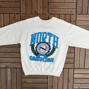 K&ouml;nnte beinhalten: Wei&szlig;es Sweatshirt mit Rundhalsausschnitt und dem Schriftzug "NORTH CAROLINA" in Blau. Das Sweatshirt hat ein rundes Emblem mit Text und einem Lorbeerkranz-Design. Das Kleidungsst&uuml;ck liegt flach auf einer Holzoberfl&auml;che.