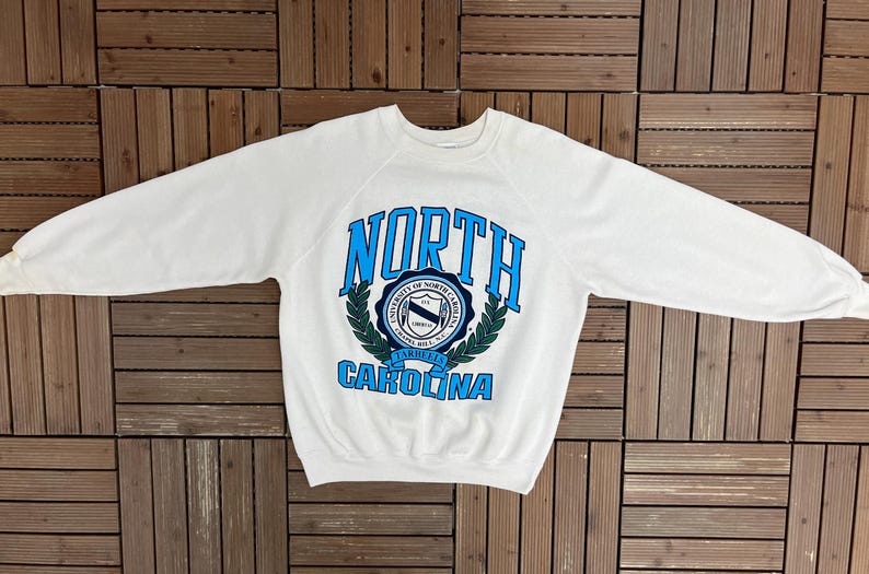 K&ouml;nnte beinhalten: Wei&szlig;es Sweatshirt mit Rundhalsausschnitt und dem Schriftzug "NORTH CAROLINA" in Blau. Das Sweatshirt hat ein rundes Emblem mit Text und einem Lorbeerkranz-Design. Das Kleidungsst&uuml;ck liegt flach auf einer Holzoberfl&auml;che.