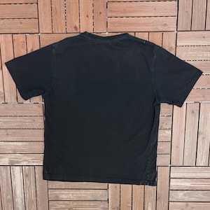 May include: A black short-sleeved t-shirt, viewed from the back, is displayed on a wooden surface. The shirt has a crew neck and a simple, classic design. The fabric appears to be soft and well-worn.