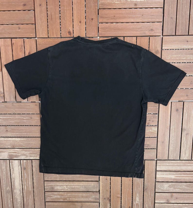 May include: A black short-sleeved t-shirt, viewed from the back, is displayed on a wooden surface. The shirt has a crew neck and a simple, classic design. The fabric appears to be soft and well-worn.