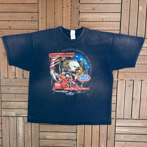 Può includere: Una t-shirt blu navy a maniche corte con una grafica raffigurante un'auto da drag racing rossa e un'aquila calva. Il design include il testo Championship Drag Racing e il logo NHRA. La maglia presenta un girocollo e una vestibilità standard.
