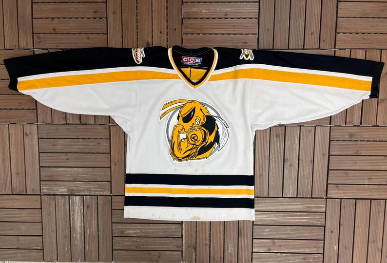 May include: White hockey jersey with black and yellow accents. The jersey features a large cartoon wasp logo on the front, with black sleeves and yellow stripes. The jersey has a V-neck collar and the CCM logo.