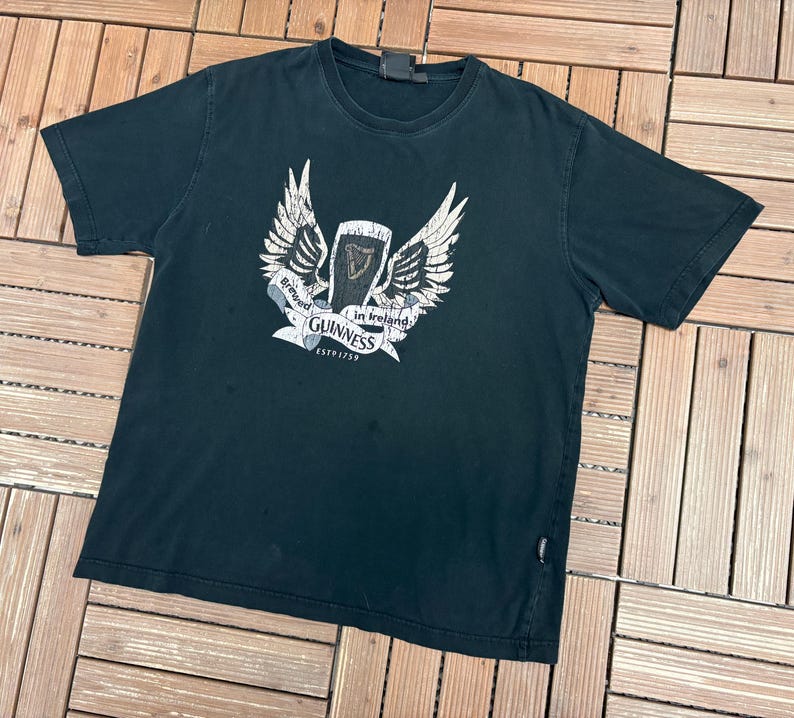 May include: Black t-shirt featuring a white graphic of a pint of Guinness with wings and the text "Brewed in Ireland Guinness ESTD 1759". The shirt is made of a soft material and has a crew neck.