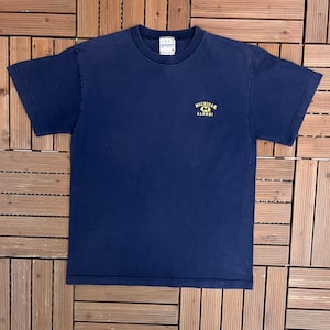 May include: A navy blue crew neck t-shirt with a small yellow and blue "Michigan Alumni" logo. The shirt is made of cotton and is displayed on a wooden surface. The t-shirt is a size medium.