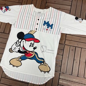 May include: A white baseball jersey with red and blue pinstripes on the upper chest, featuring a large graphic of Mickey Mouse swinging a bat. The shirt includes the letters MM, three stars, and small Mickey Mouse graphics on the sleeves.