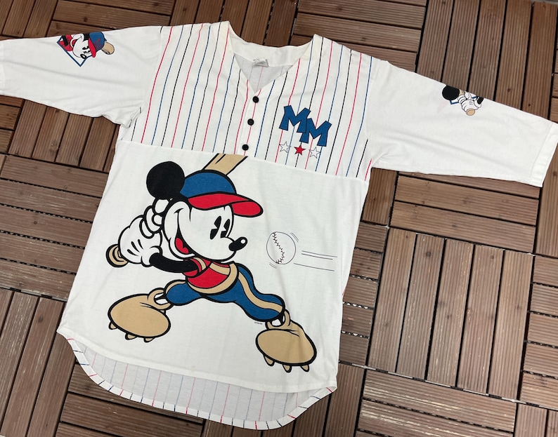May include: A white baseball jersey with red and blue pinstripes on the upper chest, featuring a large graphic of Mickey Mouse swinging a bat. The shirt includes the letters MM, three stars, and small Mickey Mouse graphics on the sleeves.
