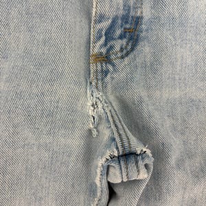 May include: Close-up of light blue denim jeans with distressed details. The image focuses on the front of the jeans, showcasing the button area and a torn section of the fabric. The stitching is visible, and the denim has a faded appearance.