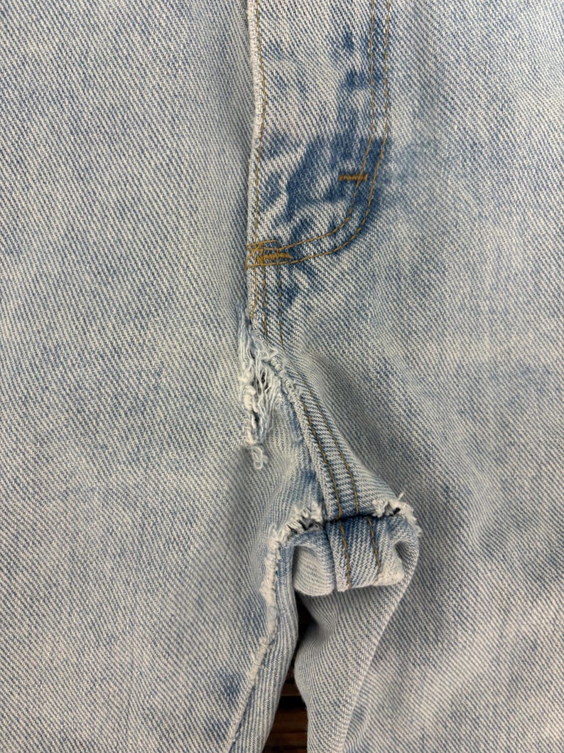 May include: Close-up of light blue denim jeans with distressed details. The image focuses on the front of the jeans, showcasing the button area and a torn section of the fabric. The stitching is visible, and the denim has a faded appearance.