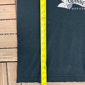 May include: Black t-shirt with a Guinness logo, featuring the text "Brewed in Guinness ESTD 1759". The shirt is approximately 29 inches long, as measured by a yellow tape measure. The shirt is made of a dark fabric.