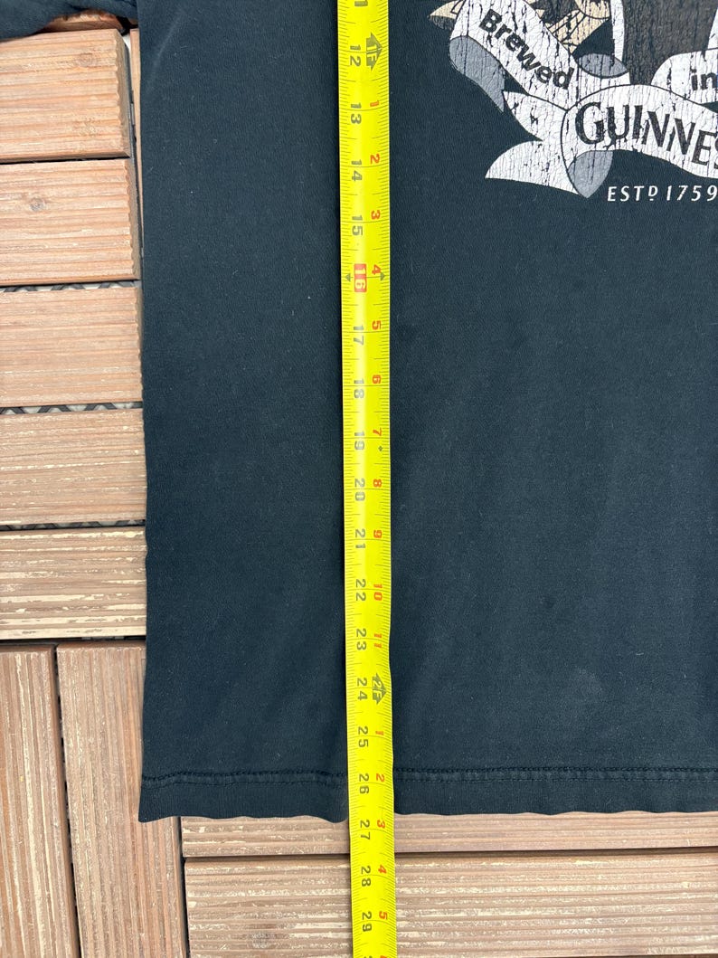 May include: Black t-shirt with a Guinness logo, featuring the text "Brewed in Guinness ESTD 1759". The shirt is approximately 29 inches long, as measured by a yellow tape measure. The shirt is made of a dark fabric.